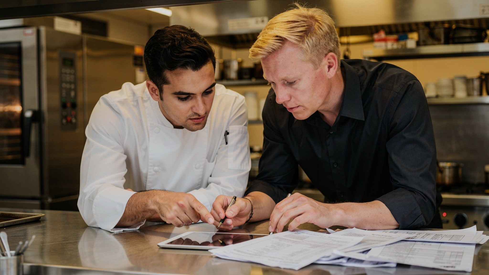 Restaurantchef en manager bekijken samen voorraadafwijkingsrapporten en tabletdata aan de keukentoonbank