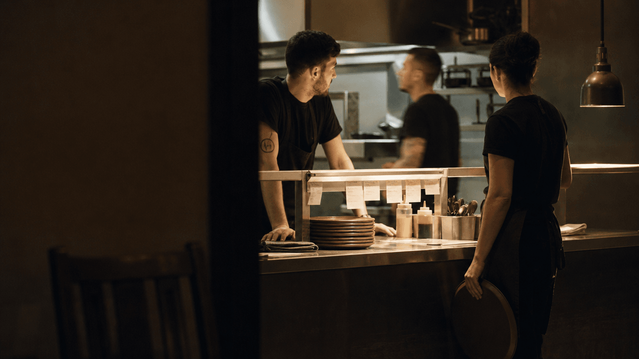 Cook and server talking across a service pass with tickets clipped above the plates