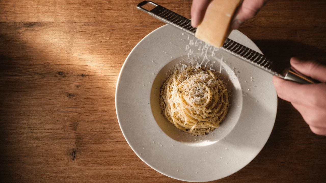 Hands grating hard cheese over a plated nest of pasta, seen from above