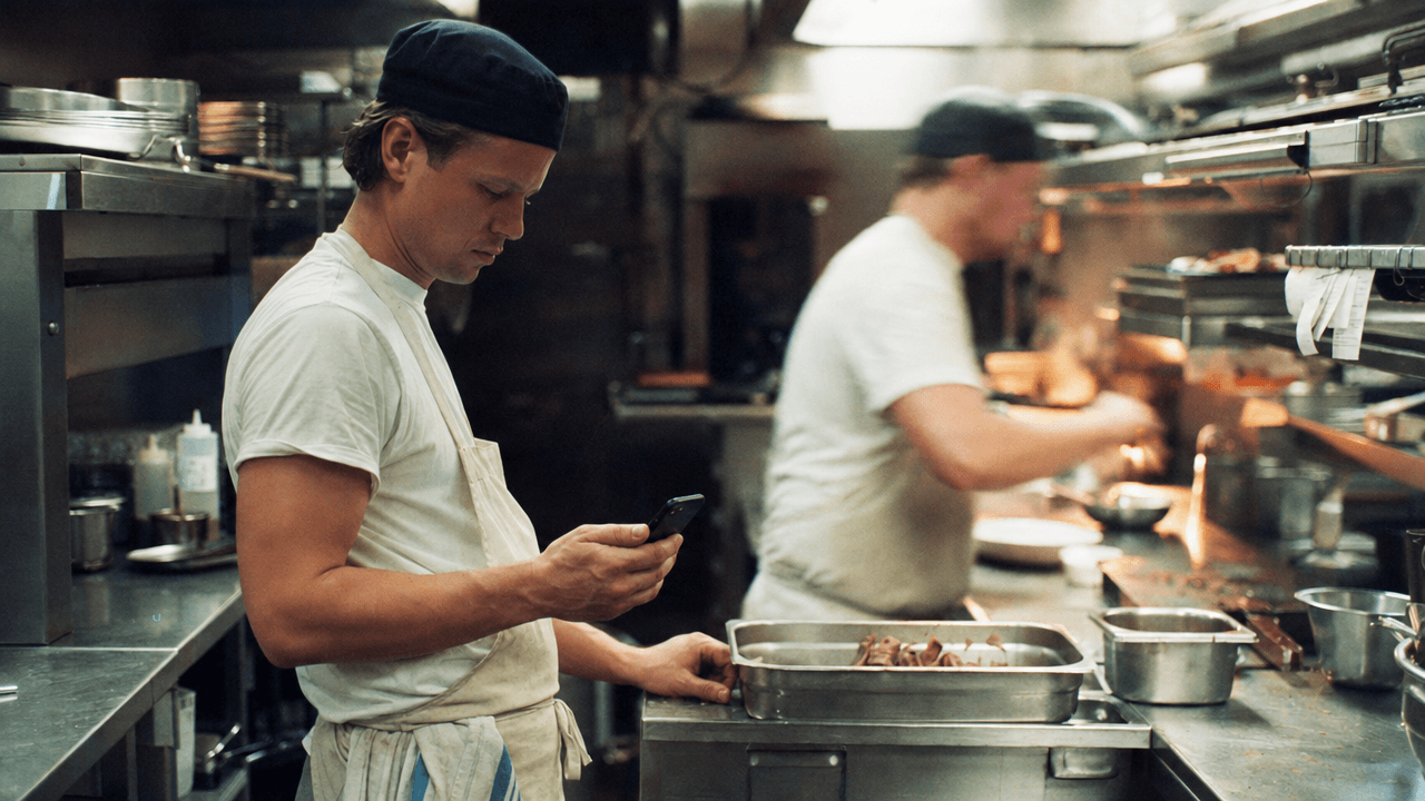Cook logging kitchen trim on a phone at the line while service continues behind